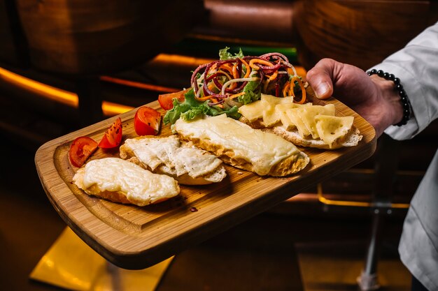 Vista lateral, um homem segura uma bandeja com torradas de queijo com fatias de tomate e salada de legumes