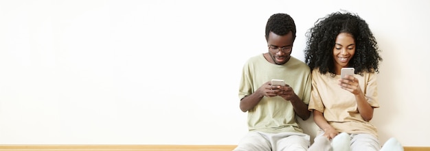 Vista panorâmica de um homem e uma mulher de pele escura, vestidos casualmente, usando telefones celulares e sorrindo, sentados no chão