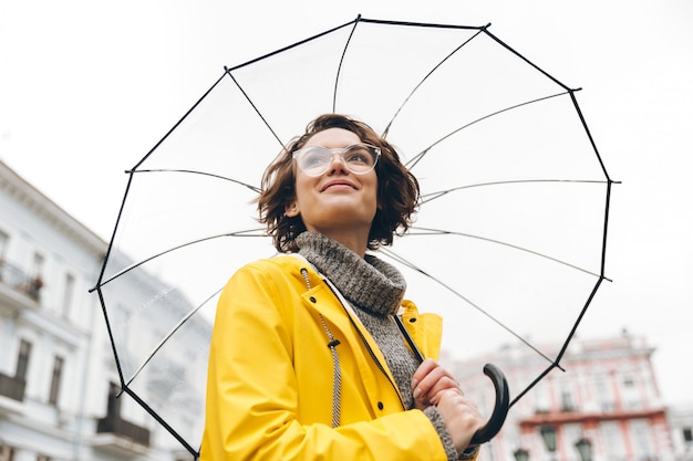 Vista por baixo da mulher positiva na capa de chuva amarela e óculos em pé na rua sob o grande guarda-chuva transparente durante o dia chuvoso cinza