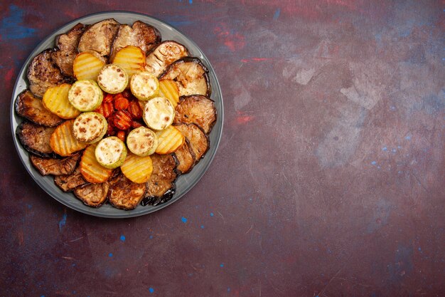 Vista superior, batatas e berinjelas assadas com legumes dentro do prato na mesa escura