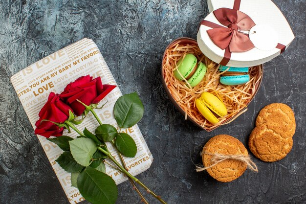 Vista superior da bela caixa de presente em forma de coração com deliciosos macarons e biscoitos de rosa vermelha em fundo escuro glacial