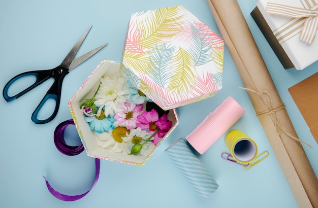 Foto grátis vista superior da caixa de presente cheia de flores de crisântemo colorido com margarida e tesoura rolos de papel e clipes de papel em fundo azul