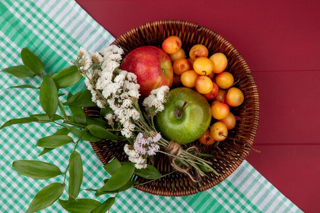 Vista superior da cereja branca com maçãs coloridas e flores em uma cesta sobre uma mesa vermelha