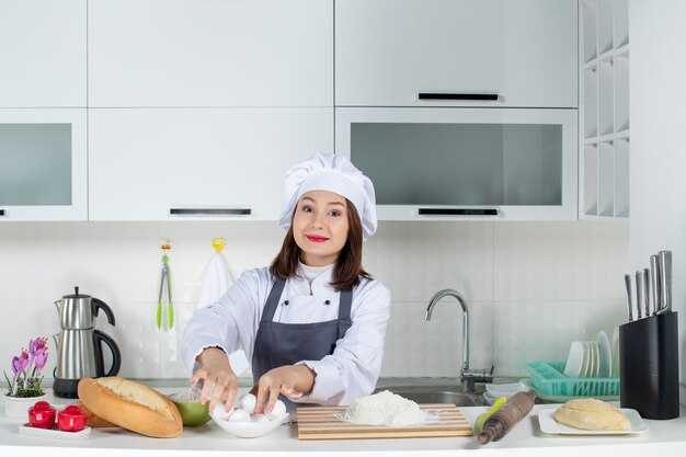 Vista superior da chef feminina de uniforme em pé atrás da mesa com uma tábua de cortar alimentos levando ovos na cozinha branca