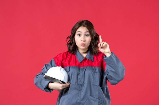 Vista superior da construtora surpresa de uniforme e segurando um capacete apontando para cima sobre um fundo vermelho isolado