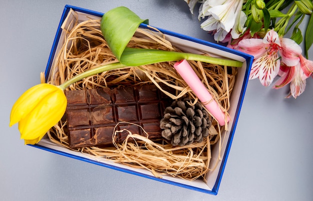 Vista superior da flor de tulipa de cor amarela com barra de chocolate escura e cone em um canudo em uma caixa de presente azul e um buquê de cores de alstroemeria na mesa branca