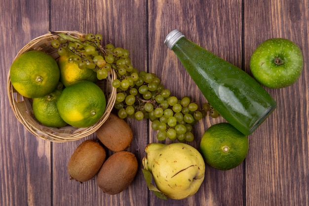 Vista superior da garrafa de suco com pêra kiwi tangerinas, maçãs e uvas em uma cesta na parede de madeira