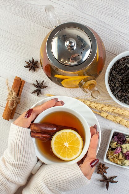 Vista superior da mão de uma mulher segurando uma xícara com chá de ervas com canela, limão e limão e os ingredientes necessários em fundo branco