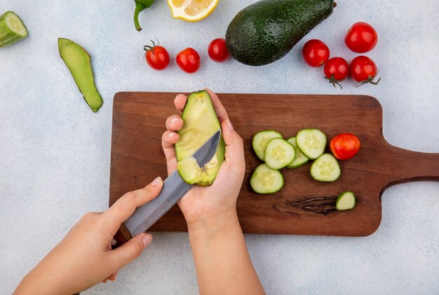 Vista superior da mão feminina segurando em uma mão o abacate e na outra a faca sobre a placa de madeira da cozinha com fatias de pepino, tomate cereja e limão isolado no branco