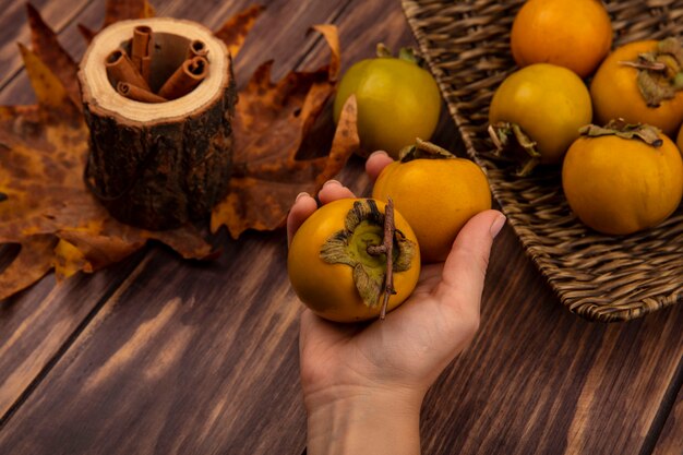 Vista superior da mão feminina segurando frutas frescas de caqui com paus de canela em uma jarra de madeira com folhas em uma mesa de madeira