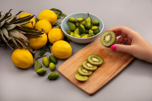 Vista superior da mão feminina segurando kiwi fresco em uma placa de cozinha de madeira com kinkans em uma tigela com limões isolados em um fundo branco