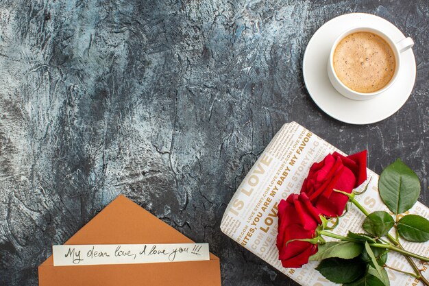 Vista superior da mão segurando rosas vermelhas em uma linda caixa de presente e um envelope de xícara de café com uma carta de amor no lado esquerdo em um fundo escuro e gelado