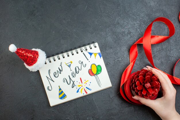 Vista superior da mão segurando um cone de conífera com fita vermelha e caderno com a escrita de ano novo e chapéu de papai noel em fundo escuro