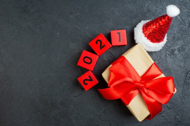 Vista superior da mão segurando um lindo presente com fita em forma de arco ao lado dos números do chapéu de Papai Noel em fundo escuro