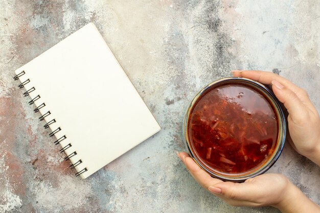 Vista superior da mão segurando um pote azul com uma deliciosa sopa borscht e um caderno espiral em um fundo colorido