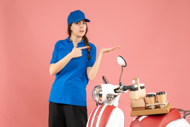 Vista superior da mensageira em pé ao lado da motocicleta com café e pequenos bolos apontando algo no lado esquerdo em um fundo de cor pastel de pêssego