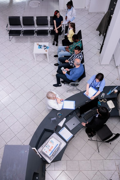 Vista superior da recepção da clínica com enfermeira profissional e médico sênior perguntando à recepcionista sobre a próxima consulta. sala de espera do hospital com diversas pessoas falando sobre oferta de folheto promocional.