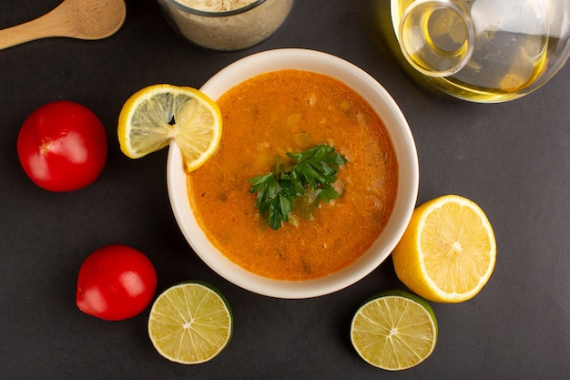 Vista superior da saborosa sopa de vegetais dentro do prato com óleo de rodelas de limão e tomate vermelho na superfície escura