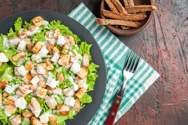 Vista superior da salada caesar em prato oval, tigela de guardanapo quadriculado verde branco com crosta seca em fundo vermelho escuro