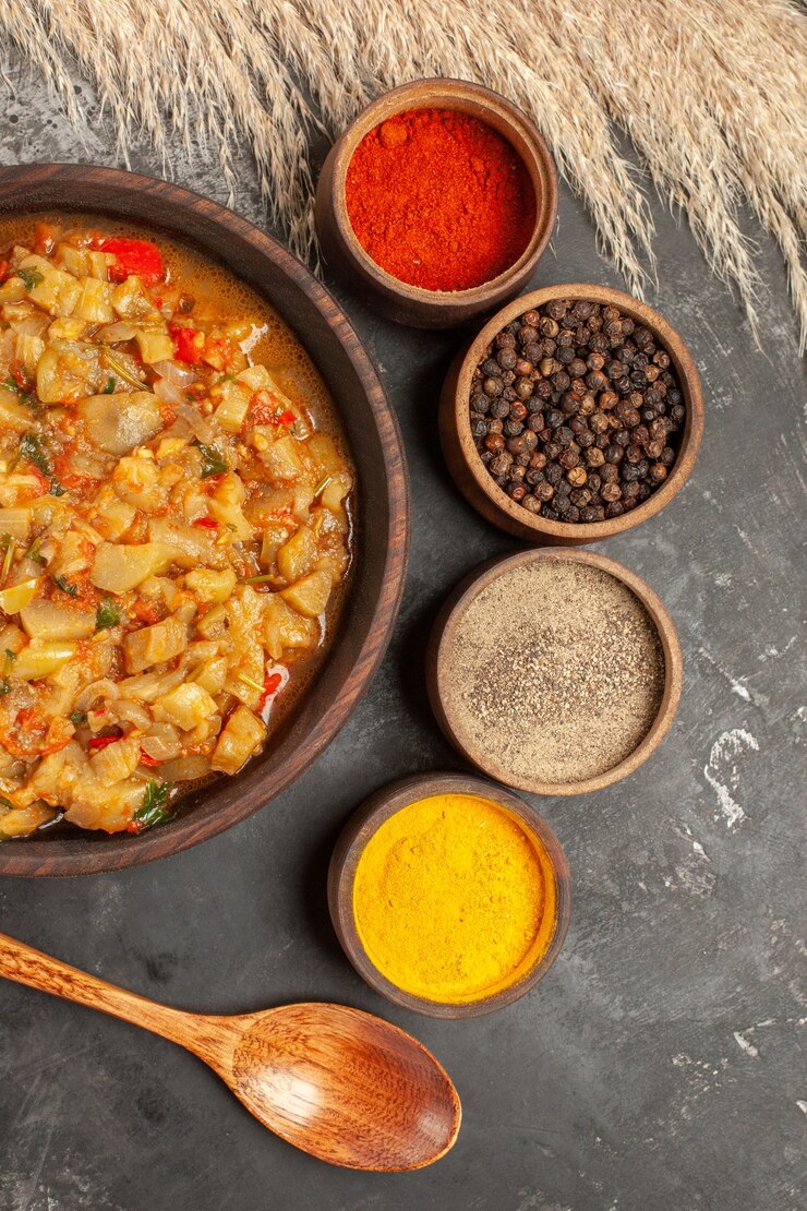 Foto grátis vista superior da salada de berinjela assada em uma tigela e diferentes especiarias em pequenas tigelas uma colher de pau na superfície escura