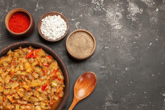 Vista superior da salada de berinjela assada em uma tigela e especiarias diferentes em tigelas, colher de pau na superfície escura