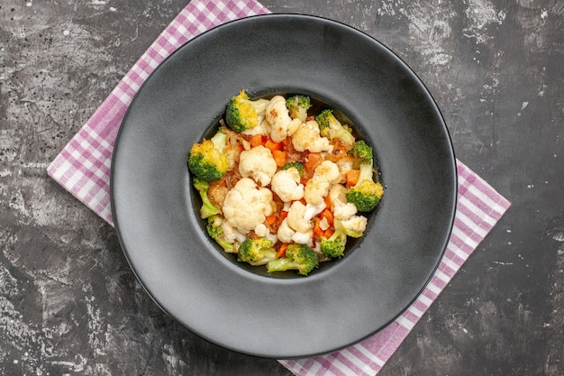 Vista superior da salada de brócolis e couve-flor em prato oval preto na bandeja de serviço cinza toalha de mesa quadriculada rosa e branca na superfície escura