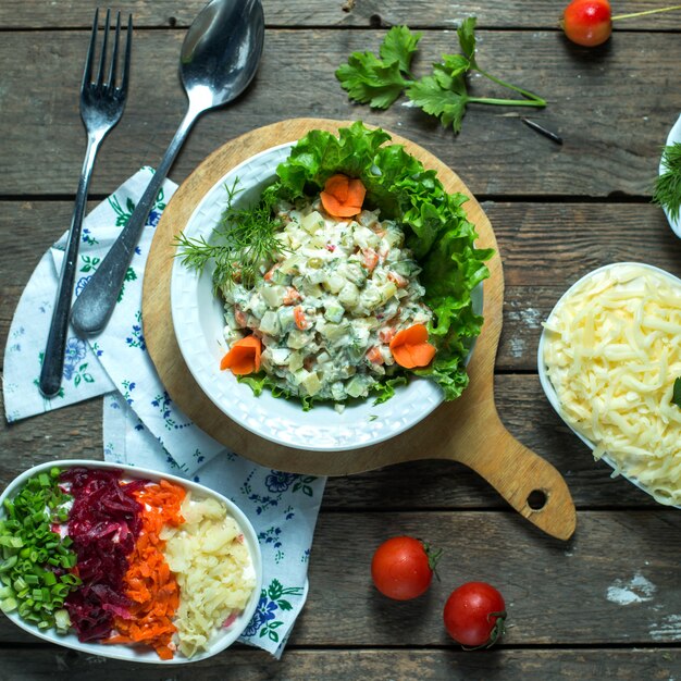 Vista superior da salada olivier russa tradicional com ervilha de frango verde e legumes em um prato branco em uma placa de madeira