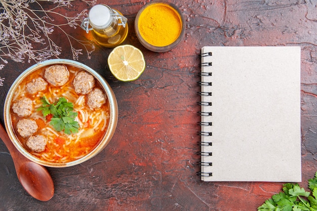 Vista superior da sopa de almôndegas com macarrão em uma tigela marrom, colher de limão, garrafa de óleo e caderno na mesa escura