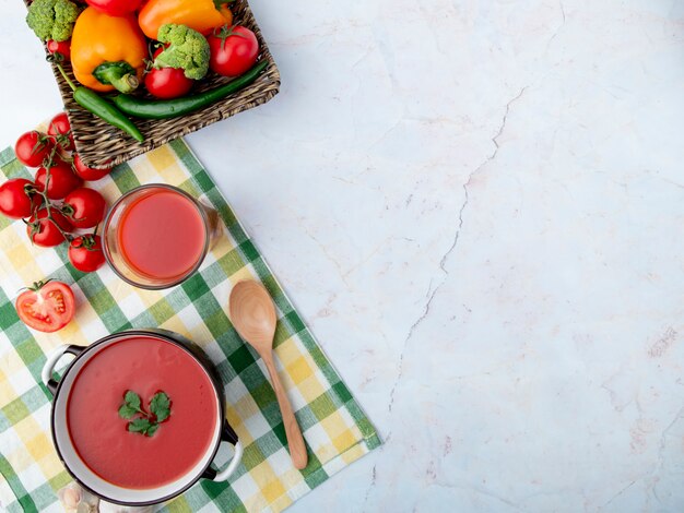 Vista superior da sopa de tomate com suco de tomate e colher e legumes no lado esquerdo na superfície branca