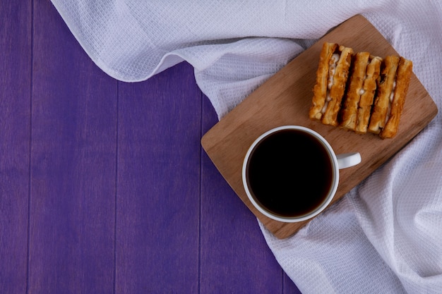 Vista superior da xícara de café e bolos na tábua no pano branco e fundo roxo