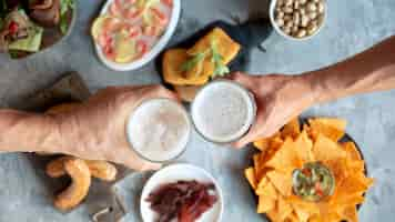 Foto grátis vista superior das duas mãos com copos de cerveja e deliciosos petiscos.