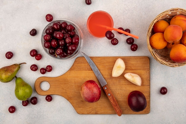 Vista superior das frutas inteiras e pêssegos cortados com faca na tábua e suco de cereja com jarra de cereja e cesta de damasco com peras no fundo branco