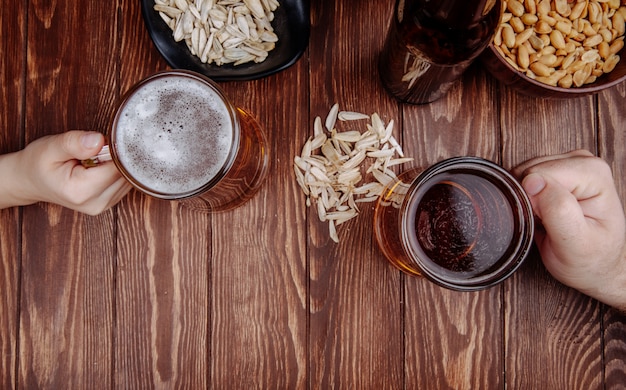 Vista superior das mãos com canecas de cerveja e salgadinhos sementes de girassol em madeira rústica