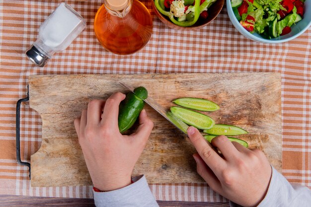 Vista superior das mãos cortando pepino com faca na tábua com salada de legumes derretida sal de manteiga na superfície do pano xadrez