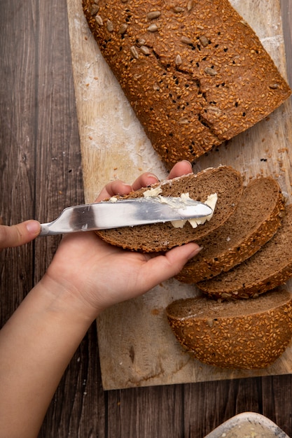 Vista superior das mãos de mulher, espalhando manteiga no pão e fatias de pão na superfície de madeira e fundo