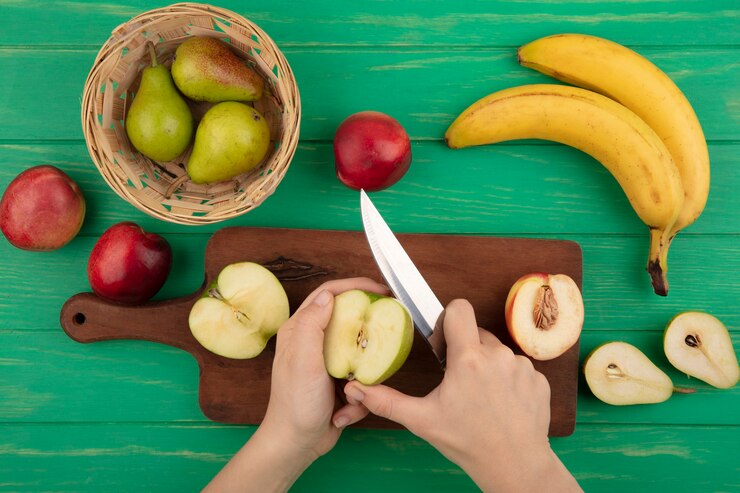 Foto grátis vista superior das mãos femininas cortando maçã com faca e meia maçã e pêssego na tábua com peras na cesta e bananas pêssegos e meia pera cortada em fundo verde