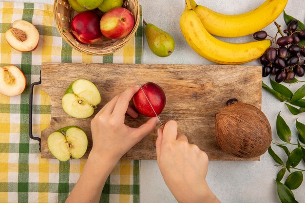 Vista superior das mãos femininas cortando pêssego com faca e metade cortada maçã e coco na tábua e cesta de maçã pêssego em pano xadrez com uvas de banana e folhas no fundo branco