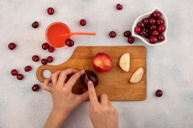 Vista superior das mãos femininas cortando pêssego com faca na tábua e suco de cereja com tigela de cereja no fundo branco
