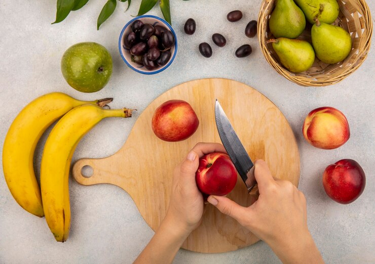Foto grátis vista superior das mãos femininas cortando pêssego com uma faca na tábua e uva pêra banana maçã com folhas no fundo branco