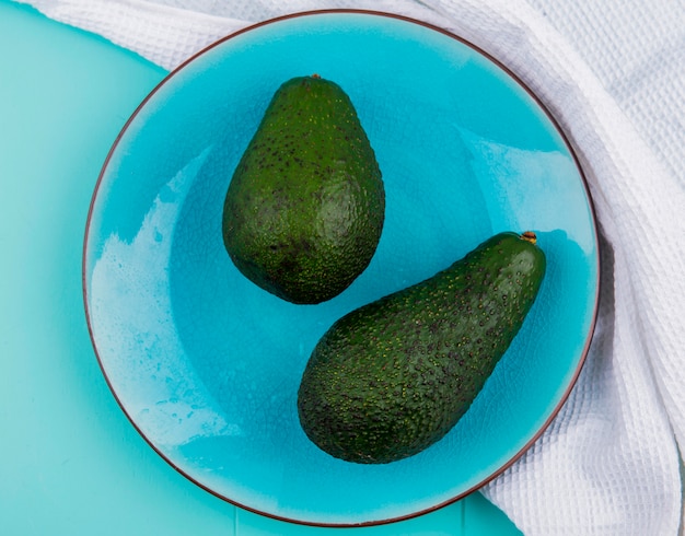 Vista superior de abacates verdes e frescos em um prato numa toalha de mesa branca na superfície azul