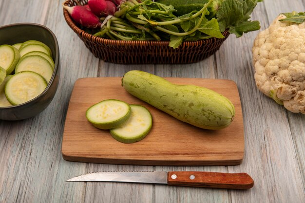 Vista superior de abobrinha verde fresca em uma placa de cozinha de madeira com uma faca com couve-flor isolada em uma superfície de madeira cinza