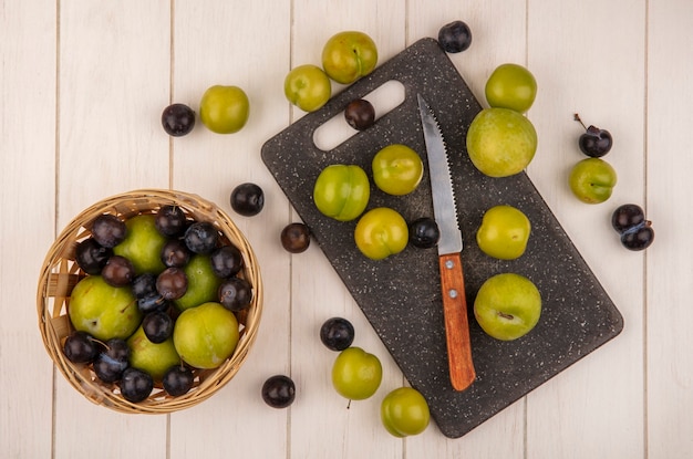 Vista superior de ameixas de cereja verdes frescas em uma tábua de cozinha com uma faca com ameixas de cereja e abrunhos em um balde em um fundo branco de madeira