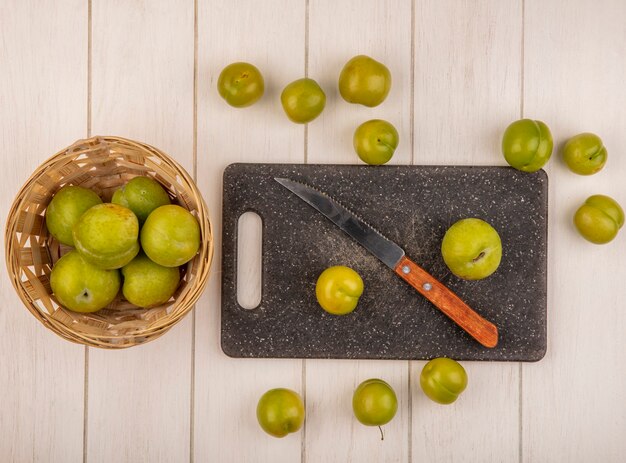 Vista superior de ameixas de cereja verdes frescas em uma tábua de cozinha com uma faca com ameixas de cereja em um balde em um fundo branco de madeira