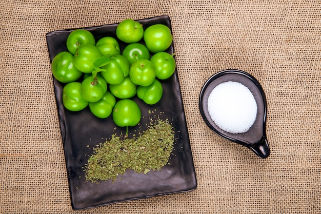 Foto grátis vista superior de ameixas verdes azedas com hortelã-pimenta seca em uma bandeja preta e sal na mesa de textura de pano de saco