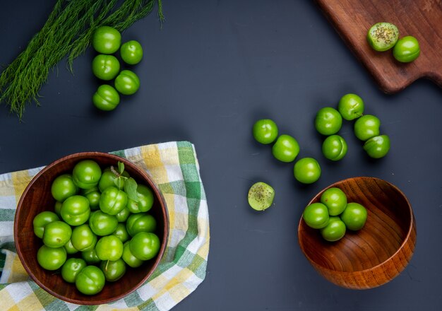 Vista superior de ameixas verdes azedas em bacias de madeira e tábua de madeira com ameixas fatiadas na mesa preta