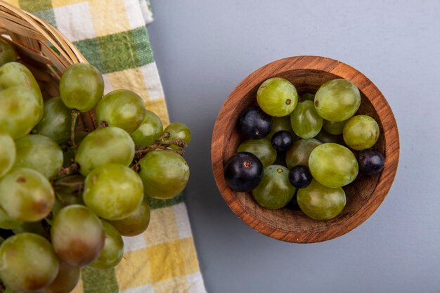 Vista superior de bagas de uva em uma tigela com uma cesta de uvas em tecido xadrez e fundo cinza