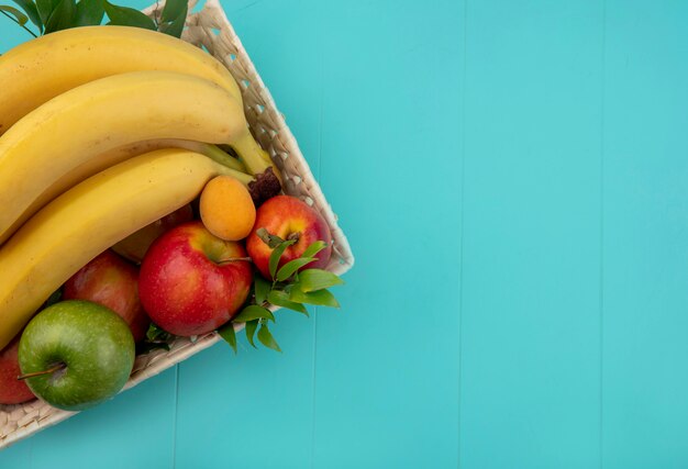 Vista superior de bananas com maçãs coloridas e pêssego em uma cesta com galhos em uma superfície turquesa