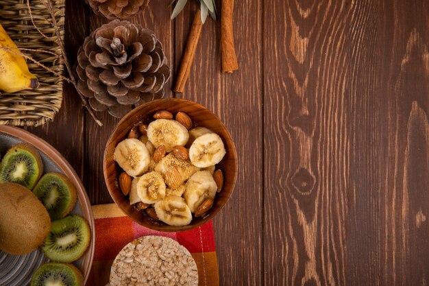 Vista superior de bananas fatiadas com amêndoa em uma tigela de madeira, fatias de kiwis em um prato e bolachas de arroz na rústica com espaço de cópia