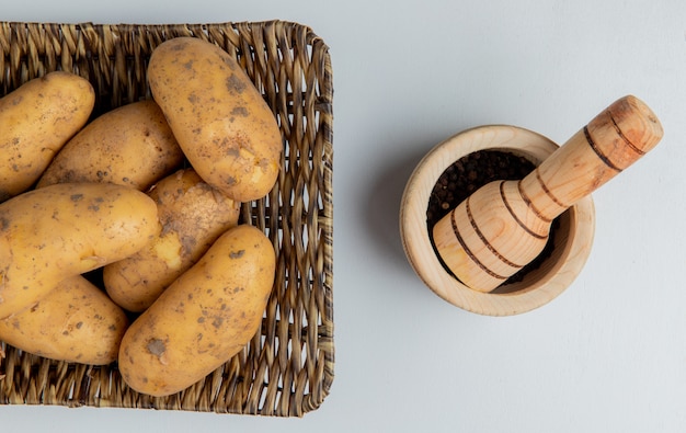 Vista superior de batatas no prato de cesto e sementes de pimenta preta no triturador de alho na superfície branca