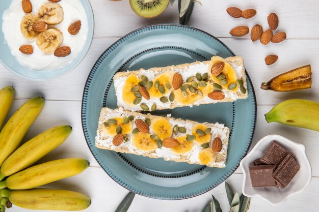 Vista superior de biscoitos crocantes com creme de queijo, fatias de banana, amêndoa e sementes de abóbora em um prato na madeira branca